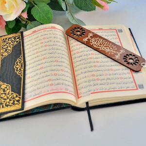 Può includere: Un libro aperto con copertina nera decorata da motivi floreali dorati, che mostra pagine con testo in arabo. Un segnalibro rettangolare in legno con disegni geometrici tagliati al laser e calligrafia araba poggia sulle pagine.