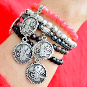 May include: A set of five bracelets with silver charms featuring intricate designs. The bracelets are made of black, gray, and pink beads.