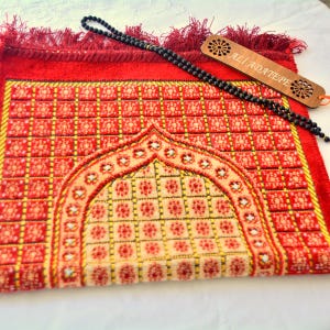 May include: A red prayer rug featuring a repeating square grid pattern and a central arch design. A string of black prayer beads rests on the rug alongside a rectangular wooden bookmark engraved with the text ALI ADATEPE and floral motifs.