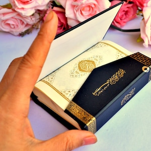 May include: A small, black, leather-bound book with gold accents and Arabic script on the cover. The book is open to reveal white pages with gold and black decorative patterns. The book is resting on a white surface.