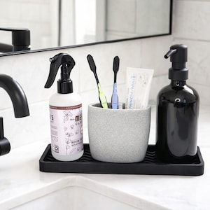 May include: A bathroom countertop arrangement featuring a black soap dispenser, a spray bottle, a toothbrush holder with toothbrushes, and a tube of toothpaste. The items are placed on a black tray, creating a cohesive and organized look.