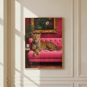 May include: A framed print featuring a leopard resting on a pink tufted sofa. The leopard has a spotted coat and is positioned in front of a painting with a gold frame. A small table with a cup and books is next to the sofa.