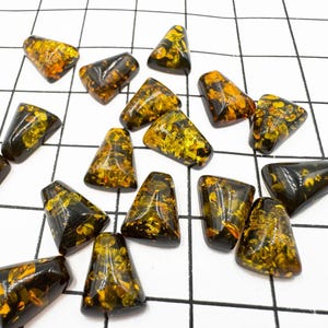 Può includere: Una collezione di cabochon di pietra preziosa ambra in varie tonalità di giallo e marrone. I cabochon hanno una forma triangolare e una superficie liscia e lucida.