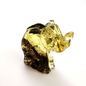 Könnte beinhalten: Eine kleine, gelbe und braune Glas-Elefantenfigur. Der Elefant sitzt mit erhobenem Rüssel.