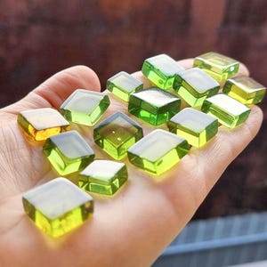 Könnte beinhalten: Eine Sammlung von quadratischen Glasfliesen in Grün und Gelb. Die Fliesen sind in einem zufälligen Muster auf einer Hand angeordnet.