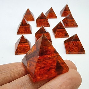 Peut inclure: Une collection de 10 pyramides en cristal d'ambre, chacune avec un motif unique et complexe de nuances brunes et orange. Les pyramides sont disposées en demi-cercle, avec une pyramide au centre tenue dans une main.