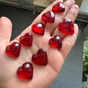 Peut inclure: Une main ouverte présente dix perles en forme de cœur, en verre rouge translucide. Les perles sont lisses et brillantes, avec des bords arrondis. La lumière naturelle se reflète sur les surfaces des cœurs.