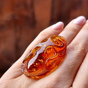 Könnte beinhalten: Nahaufnahme eines Bernsteinrings mit einer detaillierten Schnitzerei eines Drachen. Der Ring wird an einem Finger getragen.