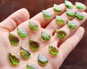 Puede incluir: Una colección de 17 colgantes de vidrio verde en forma de lágrima con destellos dorados. Los colgantes están dispuestos en una mano, mostrando su tamaño y detalle.