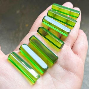 Puede incluir: Una colección de seis cuentas rectangulares de vidrio verde translúcido. Cada cuenta tiene una parte superior e inferior ligeramente redondeadas, con una superficie pulida y reflectante. Las cuentas están dispuestas en diagonal, mostrando su color y forma vibrantes.