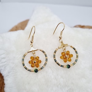 Puede incluir: Un par de pendientes de aro dorados. Cada pendiente presenta un pequeño colgante de flor de color ámbar y un anillo de pequeñas cuentas verdes oscuras y doradas. Una cuenta verde oscura está en la parte inferior de cada aro.