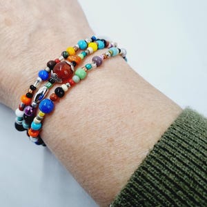 Peut inclure: Bracelets multicolores en perles avec une grosse perle de cornaline au centre. Les bracelets sont superposés au poignet d'une personne.