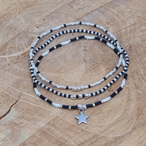 Op de afbeelding: Drie armbanden met zwarte, witte en zilveren kralen. Eén armband heeft een zilveren sterbedel. De armbanden liggen op een houten oppervlak.