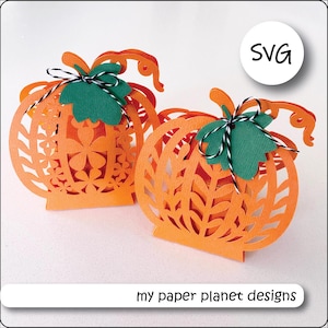 Puede incluir: Dos cajas de color naranja en forma de calabaza con intrincados diseños recortados. Las cajas tienen hojas verdes y detalles de cinta negra y blanca. Las cajas son perfectas para guardar pequeños regalos o golosinas. SVG.