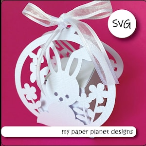 May include: White papercraft bunny rabbit favor box with a white ribbon and floral details. The box is shaped like a sphere and has a cut-out design. The text "my paper planet designs" is at the bottom of the image. The image also includes the text "SVG".