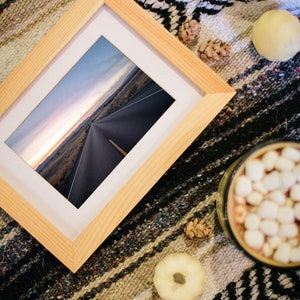 Könnte beinhalten: Ein gerahmtes Foto einer langen, geraden Straße, die zu einem fernen Horizont mit einem Sonnenuntergangshimmel führt. Der Rahmen ist aus hellem Holz und liegt auf einer gemusterten Decke mit Kürbissen und Tannenzapfen.