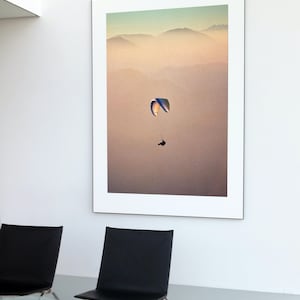Puede incluir: Una fotografía enmarcada de un parapente volando sobre una cordillera con un fondo brumoso. El cielo es de un azul suave y las montañas son de un marrón y verde apagados.