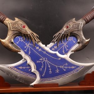 May include: Two decorative swords with blue and gold accents on a wooden stand. The swords have a sharp blade and a detailed handle with a red gem.