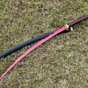 Op de afbeelding: Een rood katana-zwaard met een zwarte schede en een gouden kwast. Het zwaard ligt op een bed van groen gras.