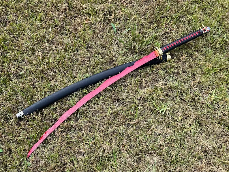 Op de afbeelding: Een rood katana-zwaard met een zwarte schede en een gouden kwast. Het zwaard ligt op een bed van groen gras.