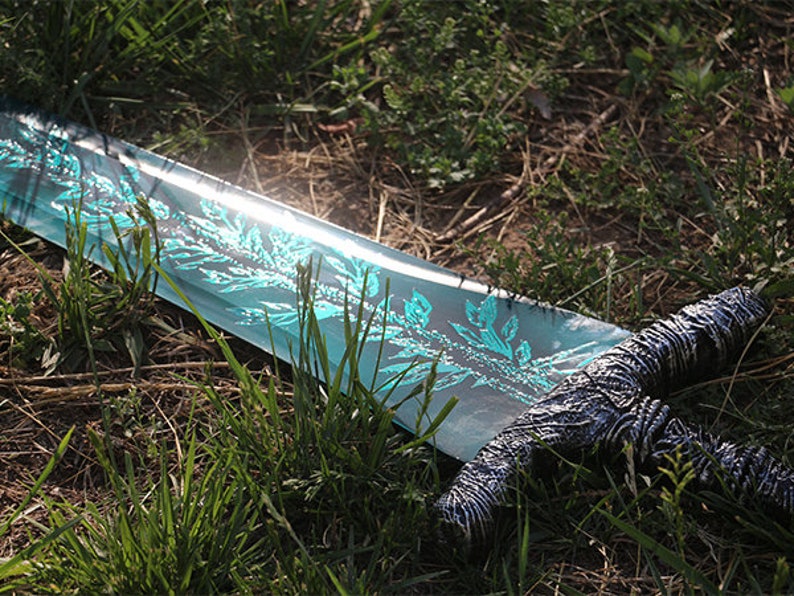 Op de afbeelding: Een blauw en zilveren zwaard met een decoratief ontwerp op het lemmet ligt op een bed van groen gras.