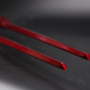 May include: A pair of red chopsticks, made of lacquered wood, are shown in a close-up view. The chopsticks are lying on a black surface.