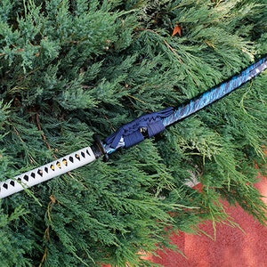 Puede incluir: Una katana negra y azul con una empuñadura blanca y dorada. La espada está sobre un lecho de follaje verde.