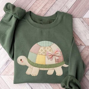 Puede incluir: Sudadera verde con un diseño de tortuga de patchwork. El caparazón de la tortuga está hecho de varias telas pastel y tiene un lazo rosa. Las mangas tienen puños fruncidos. La sudadera está doblada sobre una superficie clara de vetas de madera.