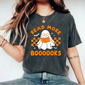 Op de afbeelding: Een grijs T-shirt met een oranje afbeelding van een geest die een bril draagt en een boek leest. De tekst "Read More BooOOOoks" staat op het T-shirt gedrukt.