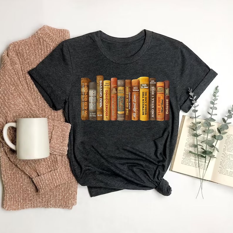 Puede incluir: Camiseta gris oscuro con un gr&aacute;fico de una fila de libros antiguos. Los t&iacute;tulos de los libros incluyen "Homegoing", "The Hate U Give" y "Between the World and Me". Los libros son de varios colores, incluyendo marr&oacute;n, amarillo y naranja. Una taza y un su&eacute;ter est&aacute;n en el fondo.