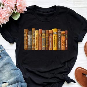 Puede incluir: Camiseta negra con un dise&ntilde;o de una fila de libros antiguos. Los t&iacute;tulos incluyen "Homegoing" y "Between the World and Me". El dise&ntilde;o tiene un tema literario.