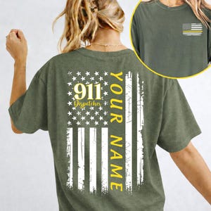 Könnte beinhalten: Olivgrünes T-Shirt mit einem verblassten amerikanischen Flaggen-Design. Der Text "911 Dispatchers" und "Your Name" sind gelb. Eine kleinere Flagge mit gelbem Streifen befindet sich auf der Brust.