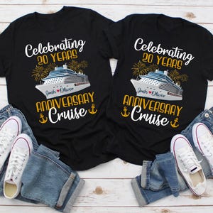 May include: Two black t-shirts with a white cruise ship graphic and gold text that reads "Celebrating 20 Years" and "Anniversary Cruise". The shirts are laid out on a wooden surface with blue jeans and white sneakers.