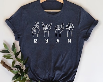 May include: A navy blue t-shirt with the name "RYAN" spelled out in British Sign Language. The letters are white and outlined in black.