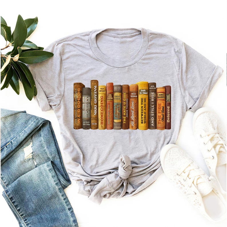 Puede incluir: Camiseta gris claro con un dise&ntilde;o de una fila de libros antiguos. Los lomos de los libros son de varios colores, con t&iacute;tulos como "The Hate U Give" y "Beloved". La camiseta se muestra con vaqueros azules y zapatillas blancas.