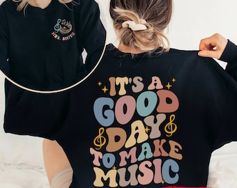 May include: Black sweatshirt with the text "It's a good day to make music" in colorful retro font. The text is surrounded by musical notes and stars. The sweatshirt can be customized.