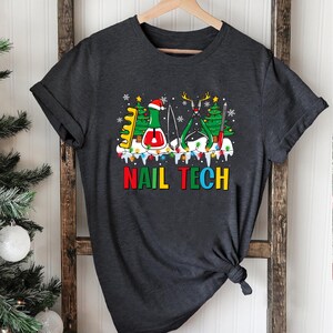 May include: A dark gray t-shirt with a Christmas-themed design featuring nail tech tools, a Christmas tree, and the text "NAIL TECH".