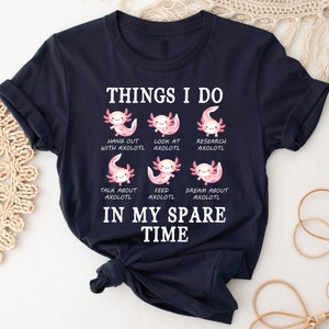 Könnte beinhalten: Ein dunkelblaues T-Shirt mit einer weißen Grafik von sechs Axolotln und dem Text "Things I do in my spare time". Die Axolotl sind rosa und haben verschiedene Gesichtsausdrücke. Der Text unter jedem Axolotl beschreibt eine Aktivität, die mit Axolotln zusammenhängt, wie z. B. "mit Axolotl abhängen", "Axolotl ansehen", "Axolotl erforschen", "über Axolotl sprechen", "Axolotl füttern" und "von Axolotl träumen".