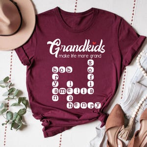 Op de afbeelding: Bordeauxkleurig T-shirt met witte tekst die "Grandkids make life more grand" zegt en een kruiswoordpuzzel met de namen Bob, Amelia, Henry en Sofi.