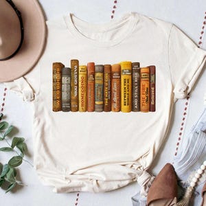 Puede incluir: Camiseta color crema con un dise&ntilde;o de lomos de libros antiguos. Los t&iacute;tulos incluyen "Homegoing", "The Hate U Give" y "Beloved". El dise&ntilde;o est&aacute; impreso en una camiseta suave e informal, ideal para los amantes de los libros.