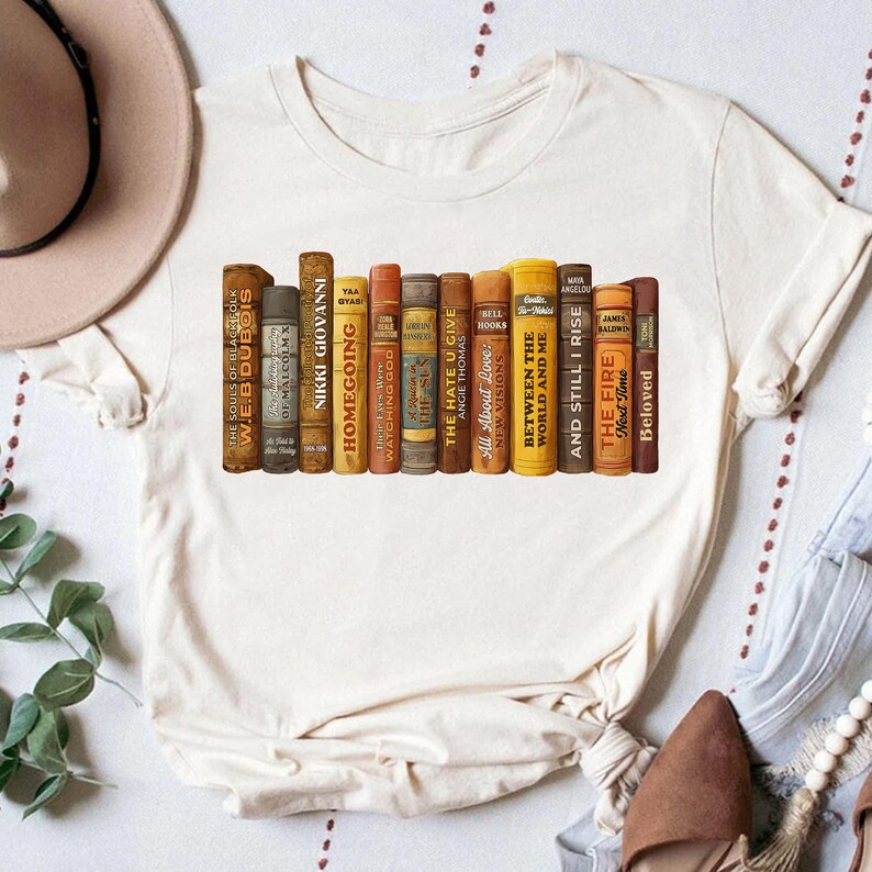 Puede incluir: Camiseta color crema con un dise&ntilde;o de lomos de libros antiguos. Los t&iacute;tulos incluyen "Homegoing", "The Hate U Give" y "Beloved". El dise&ntilde;o est&aacute; impreso en una camiseta suave e informal, ideal para los amantes de los libros.