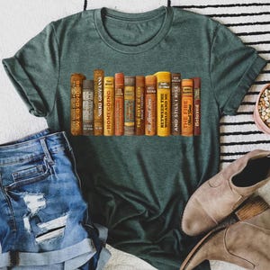 Puede incluir: Camiseta verde con un gr&aacute;fico de una fila de libros, con t&iacute;tulos como "Homegoing" y "The Hate U Give". Los libros son de varios colores, incluyendo marr&oacute;n, amarillo y naranja. La camiseta se combina con pantalones cortos de mezclilla y botas de gamuza marr&oacute;n.