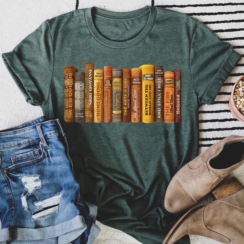 Puede incluir: Camiseta verde con un gr&aacute;fico de una fila de libros, con t&iacute;tulos como "Homegoing" y "The Hate U Give". Los libros son de varios colores, incluyendo marr&oacute;n, amarillo y naranja. La camiseta se combina con pantalones cortos de mezclilla y botas de gamuza marr&oacute;n.
