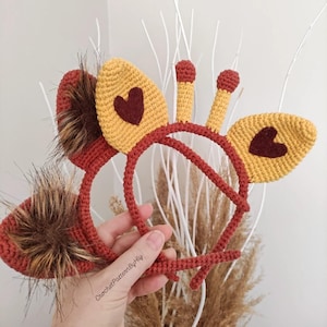 Puede incluir: Diadema de orejas de jirafa de ganchillo con orejas amarillas, detalles de corazón marrón y una diadema roja. La diadema presenta detalles de piel sintética y está diseñada para disfraces o cosplay. El texto "CrochetPatternByHly" es visible.