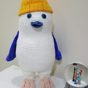 May include: A handmade, plush penguin doll with a white body, blue wings, and pink feet. It wears a yellow knitted hat with a pom-pom. The penguin has large blue eyes and a blue beak. A snow globe is in the background.