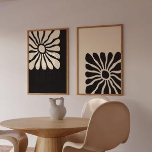 May include: Two framed black and white abstract flower prints with a cream background. The prints are hanging on a white wall above a wooden table with a white ceramic pitcher.
