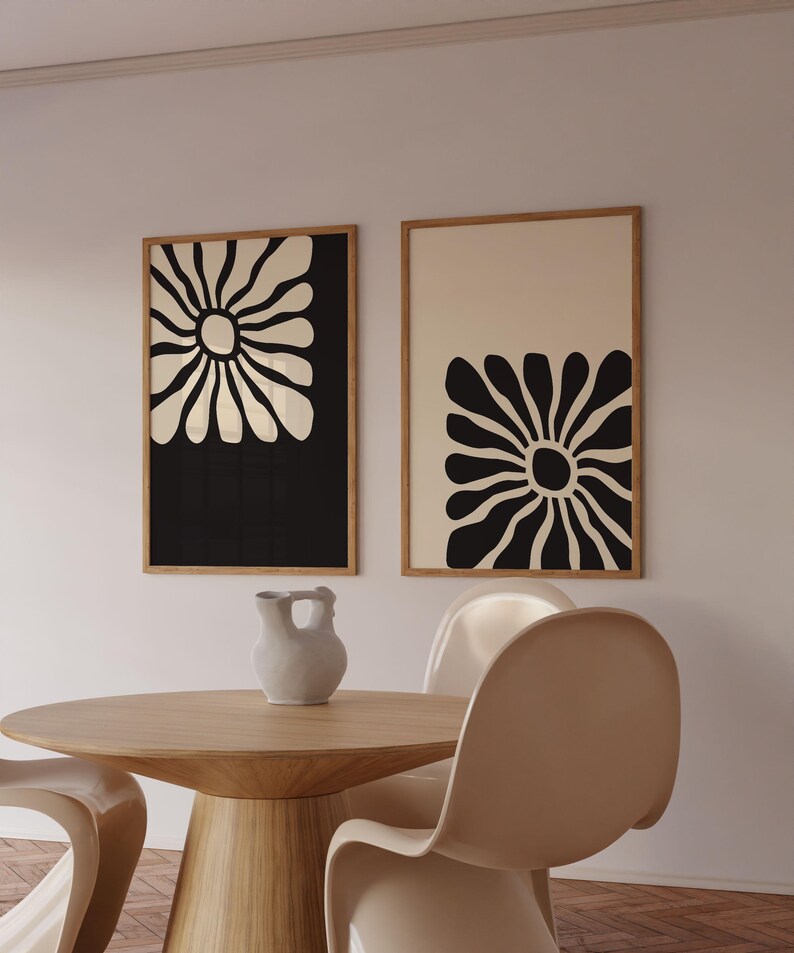 May include: Two framed black and white abstract flower prints with a cream background. The prints are hanging on a white wall above a wooden table with a white ceramic pitcher.