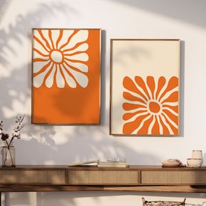 May include: Two framed prints with orange and white abstract floral designs. The prints are hung on a white wall above a wooden console table.
