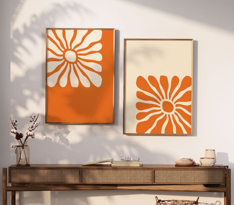 May include: Two framed prints with orange and white abstract floral designs. The prints are hung on a white wall above a wooden console table.