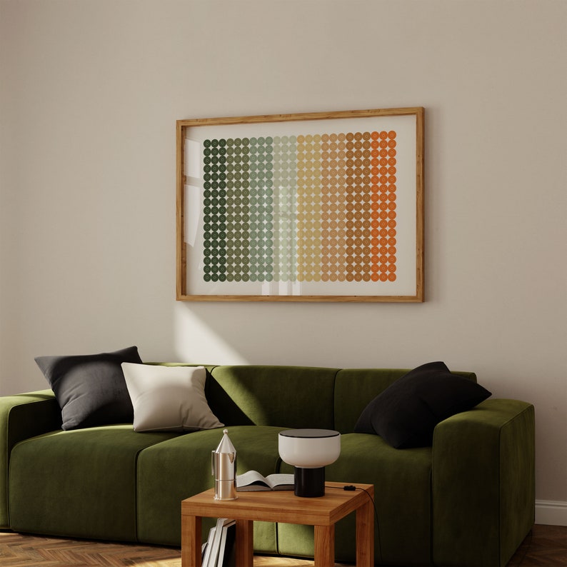 Op de afbeelding: Een groene fluwelen bank met zwarte en witte kussens staat voor een beige muur met een ingelijst abstract print van een geometrisch patroon in tinten groen, bruin en oranje. Een houten bijzettafel met een lamp en een boek staat voor de bank.